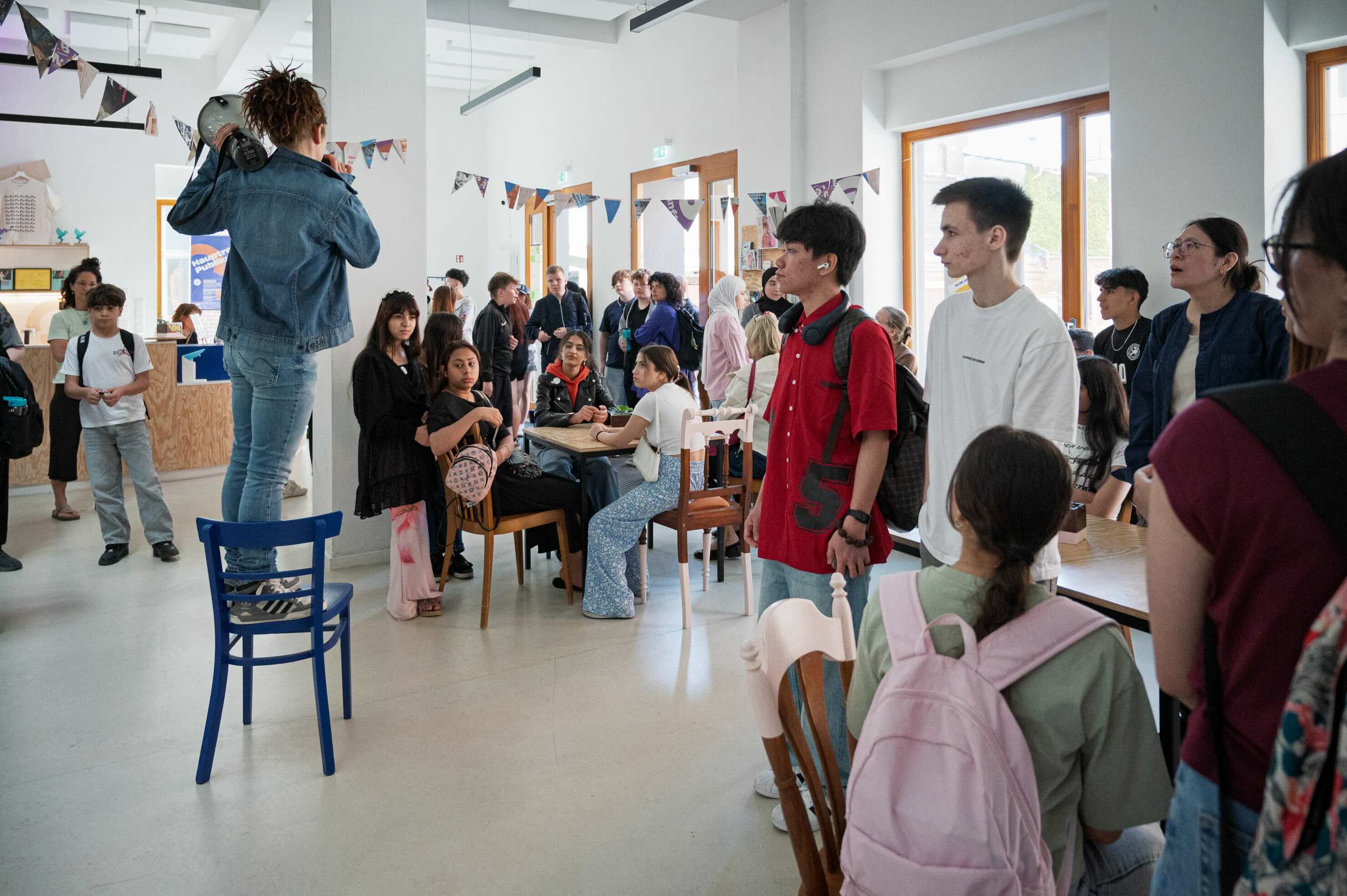 Eine Schauspielerin steht im Foyer von Theater Strahl auf einem Stuhl und macht eine Ansage. Um sie herum viele Schüler*innen.